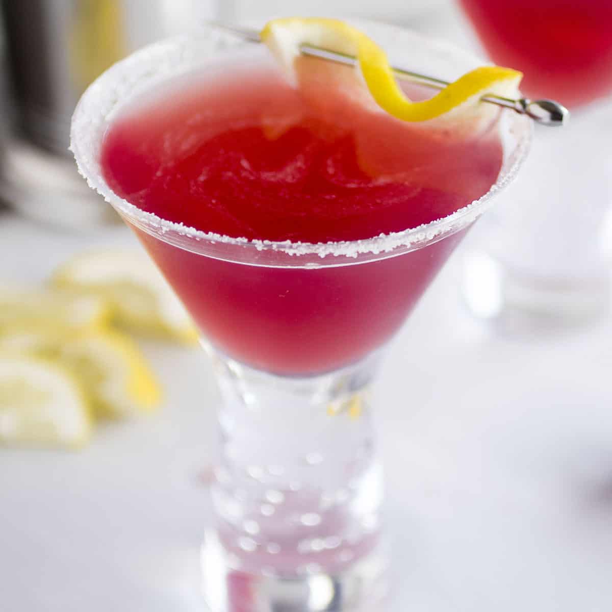 Red drink in a martini glass with a skewer of lemon peel on top.