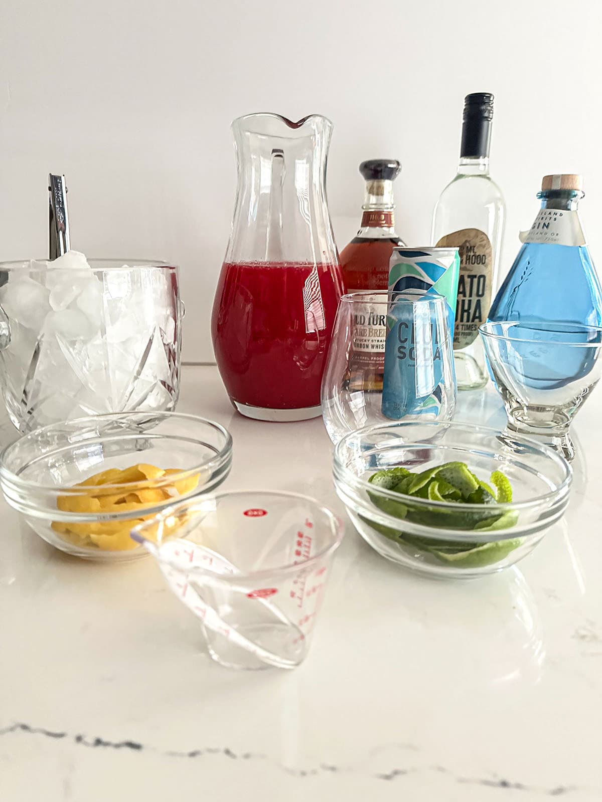 A table with a bar set-up - ice, citrus peels, glasses, spirits, mixers, and a pitcher of batch cranberry-citrus juice.