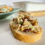 Warm jalapeño, bacon, date dip on a piece of baguette with a plate of dip and bread and the casserole dish on a table.