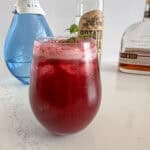 Cocktail glass with blueberry mint mule beverage and individual bottles of gin, vodka, and whiskey.